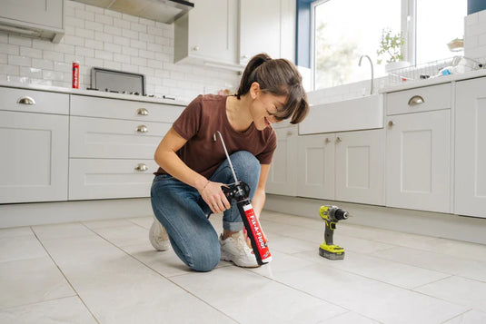 Girl using a caulking gun and fixafloor adhesive to apply sealant between floor tiles, with a cordless drill on the floor nearby.