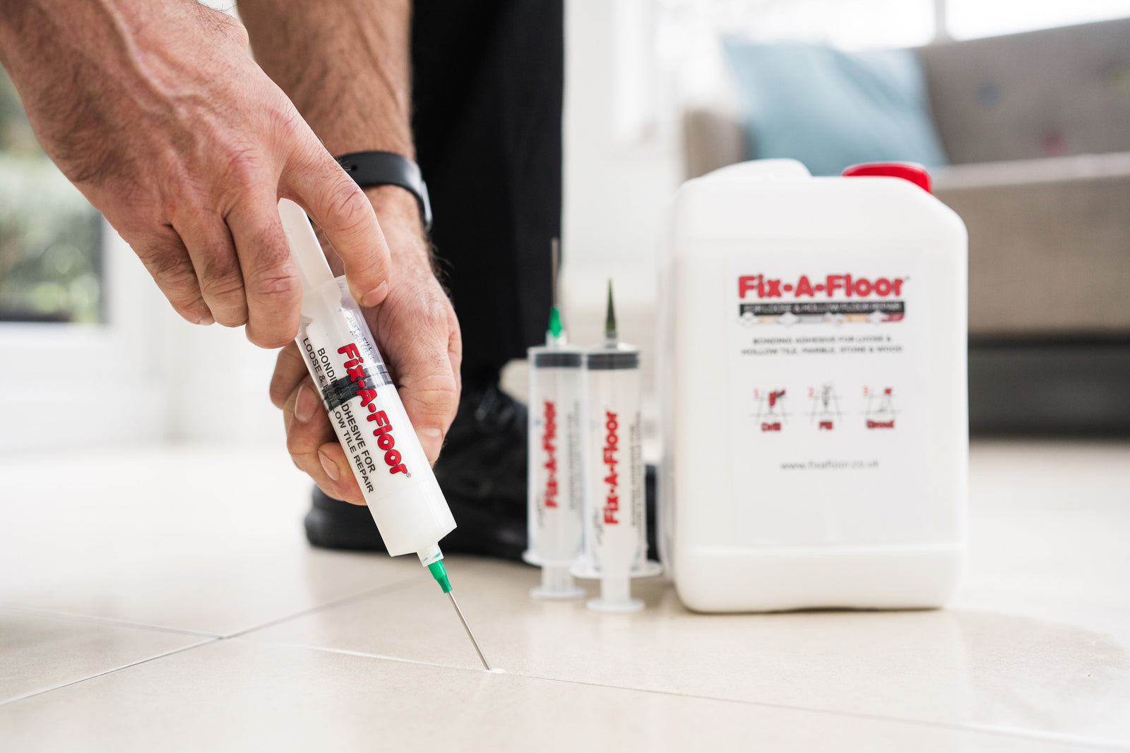 Person injecting Fix-A-Floor bonding adhesive into a tile floor grout line using a syringe applicator, with spare syringe kits and a bonding adhesive jug in the background